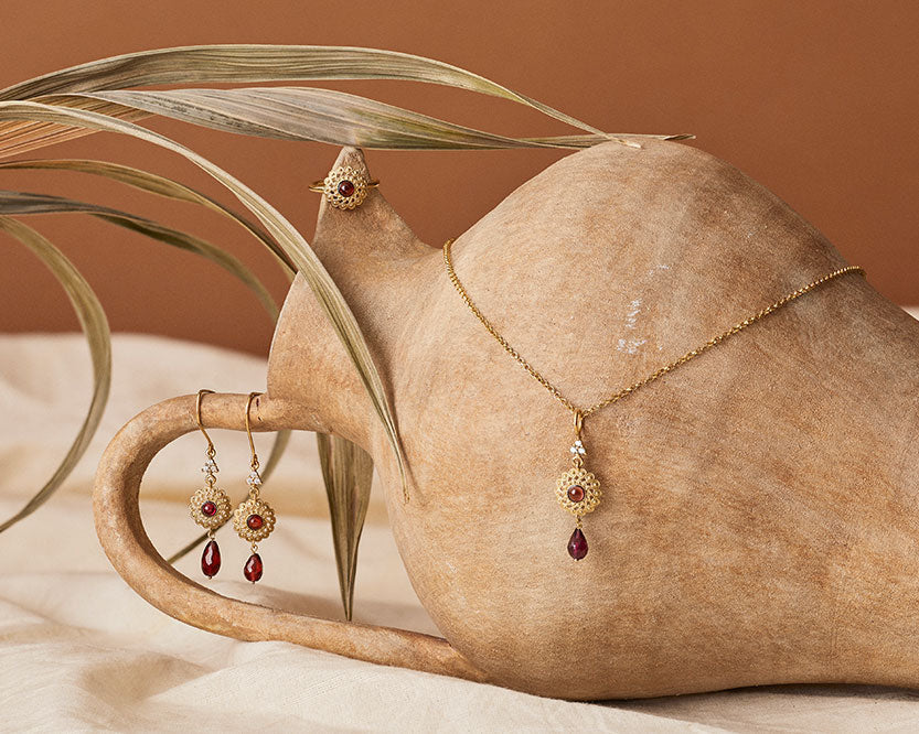 Gold-plated jewelry set with garnet — ring, earrings and necklace — styled on a ceramic vase with dried palm leaves, part of Vintouch Gifts Under €150 collection.